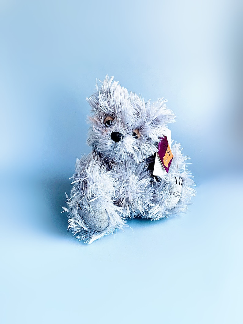 May include: A gray plush teddy bear with a tag attached to its ear. The bear is sitting on a light blue background.