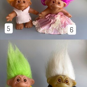 May include: Four troll dolls, two with pink and white dresses and two with yellow and black outfits. The dolls have different colored hair, including orange, pink, green, and white.