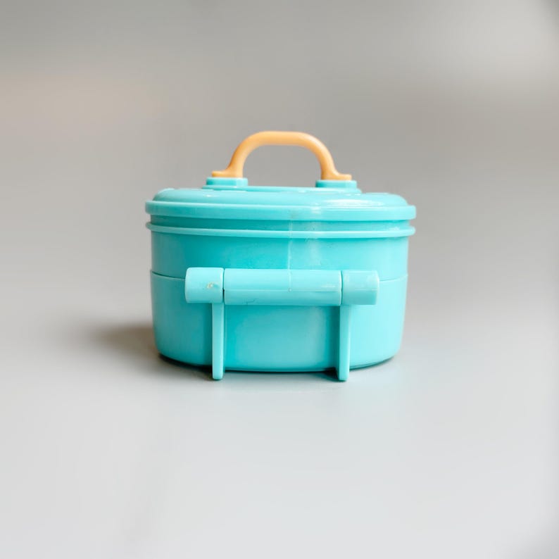 May include: A small, turquoise plastic container with a rounded shape and a light orange handle. The container has a lid and two turquoise handles on the sides. The background is a solid, neutral gray.