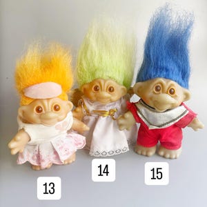 May include: Three troll dolls with different colored hair. The troll on the left has orange hair and is wearing a white dress with pink accents. The middle troll has green hair and is wearing a white dress with gold accents. The troll on the right has blue hair and is wearing a red shirt and pants with gold accents.