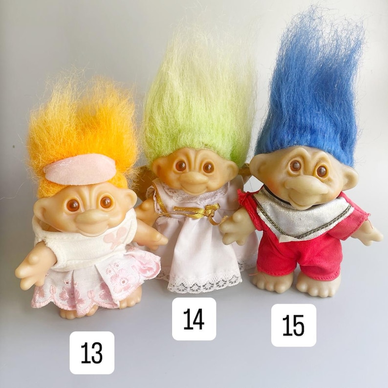 May include: Three troll dolls with different colored hair. The troll on the left has orange hair and is wearing a white dress with pink accents. The middle troll has green hair and is wearing a white dress with gold accents. The troll on the right has blue hair and is wearing a red shirt and pants with gold accents.
