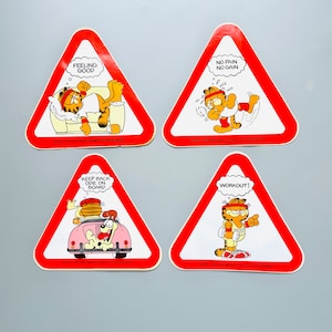 Puede incluir: Cuatro se&ntilde;ales de advertencia triangulares rojas con bordes blancos e ilustraciones de dibujos animados de Garfield el gato. Las se&ntilde;ales muestran el texto "Feeling Good", "No Pain No Gain", "Keep Back Odie On Board" y "Workout!"