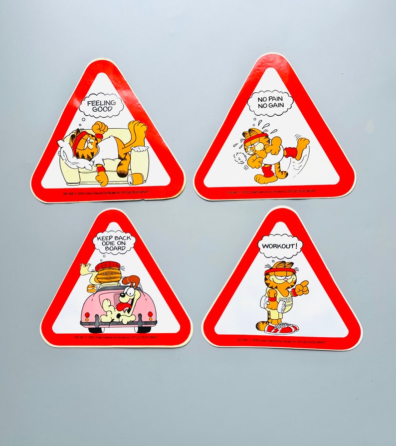 Puede incluir: Cuatro se&ntilde;ales de advertencia triangulares rojas con bordes blancos e ilustraciones de dibujos animados de Garfield el gato. Las se&ntilde;ales muestran el texto "Feeling Good", "No Pain No Gain", "Keep Back Odie On Board" y "Workout!"
