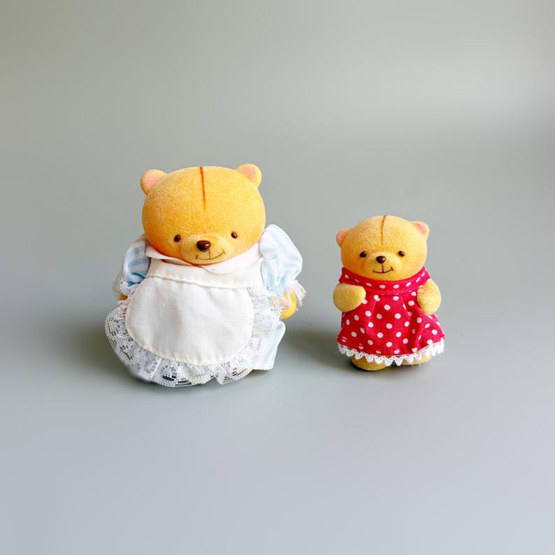 May include: Two yellow bear figurines. One bear wears a blue and white dress with a white apron. The other bear wears a red dress with white polka dots. Both bears have small, black eyes and brown noses.