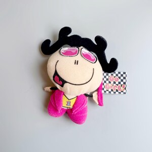 Può includere: Bambola di peluche con un viso sorridente, capelli neri ricci e occhiali da sole rosa. La bambola indossa una giacca rosa sopra una maglietta gialla con il numero 1. Un'etichetta con le parole "Go Girl!" è attaccata.