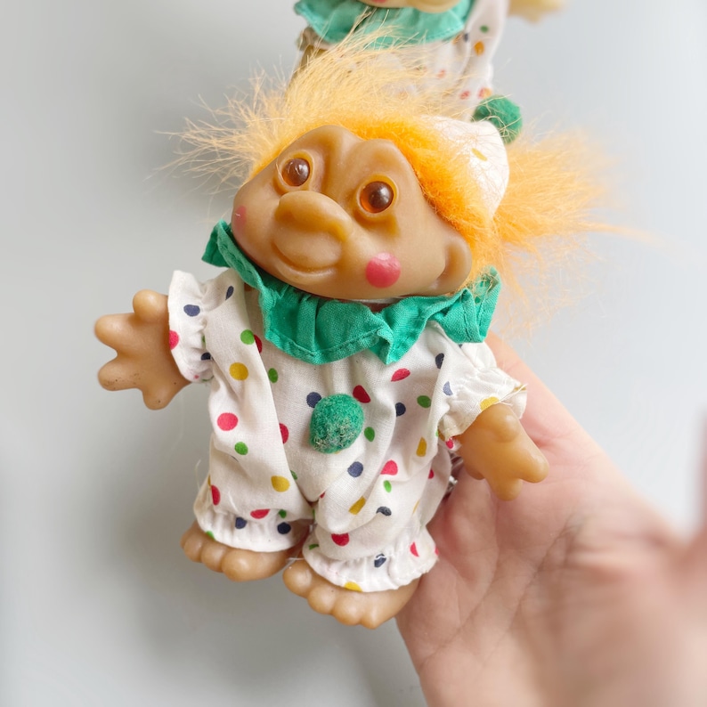 Pu&ograve; includere: Un piccolo troll con i capelli arancioni che indossa un costume da clown bianco e verde con pois colorati.