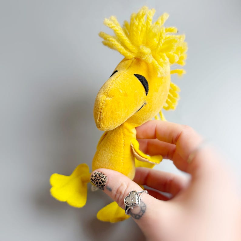 Puede incluir: Un juguete de peluche amarillo con ojos negros y un pico sonriente. El p&aacute;jaro tiene un mech&oacute;n de pelo amarillo en la cabeza y pies amarillos. El juguete se sostiene en una mano sobre un fondo gris.
