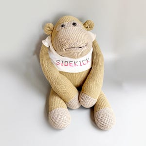 Può includere: Un peluche a forma di scimmia lavorato a maglia color marrone chiaro con una maglietta color crema che riporta la parola SIDEKICK ricamata in filo rosa. La scimmia ha occhi scuri, orecchie arrotondate e dettagli color sabbia su mani e piedi.