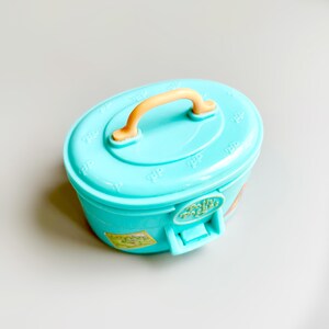 May include: A light blue plastic toy container with a peach-colored handle. The container has a rounded rectangular shape with a lid and a latch. The lid has embossed details. A sticker is visible on the side.