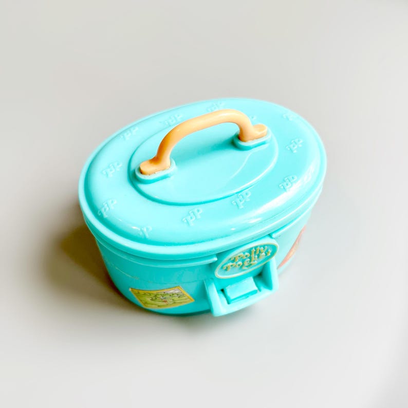 May include: A light blue plastic toy container with a peach-colored handle. The container has a rounded rectangular shape with a lid and a latch. The lid has embossed details. A sticker is visible on the side.