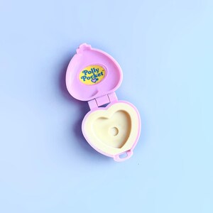 Puede incluir: Un compacto de Polly Pocket en forma de coraz&oacute;n rosa con un interior amarillo y una pegatina amarilla con las palabras "Polly Pocket" en azul.