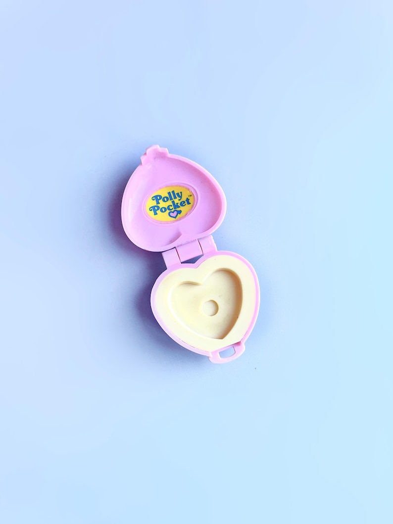 Puede incluir: Un compacto de Polly Pocket en forma de coraz&oacute;n rosa con un interior amarillo y una pegatina amarilla con las palabras "Polly Pocket" en azul.
