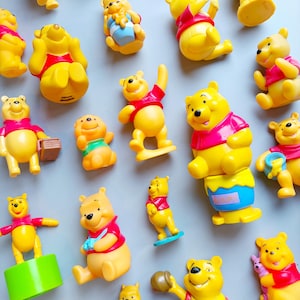 May include: A collection of yellow Winnie the Pooh figurines in various poses, wearing red shirts. Some figurines are holding objects like a book or a honey pot. The figurines are scattered on a light gray surface.