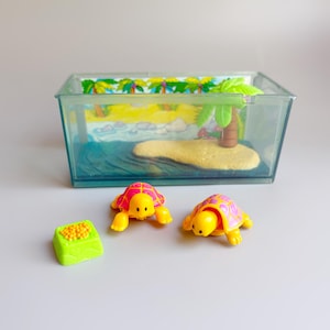 May include: A miniature toy playset featuring two yellow turtles with pink and purple patterned shells. A green food dish and a clear plastic aquarium with a painted tropical island scene are also included. The scene includes a sandy beach, ocean, and palm trees.