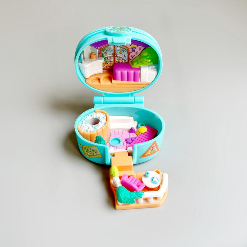 May include: A vintage, open, teal-colored Polly Pocket playset. The playset features a bedroom, a hot tub, and a small outdoor area. The interior includes a bed, a dresser, and a window. The playset is made of plastic and has a hinged design.