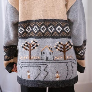 Può includere: Un maglione beige, grigio e marrone con scollo tondo. Il maglione presenta dettagli ricamati di alberi, una casa e motivi geometrici. Le maniche sono grigie e lo scollo è grigio scuro. Piccole forme ricamate sono sparse sulla sezione superiore beige.