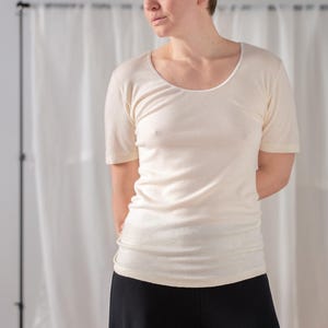 Könnte beinhalten: Ein cremefarbenes T-Shirt mit kurzen Ärmeln und Rundhalsausschnitt, kombiniert mit einer schwarzen Hose. Das Shirt hat eine taillierte Silhouette. Das Model steht vor einem weißen Vorhang.