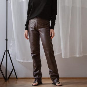 May include: A person wearing a black turtleneck sweater and brown leather pants. The outfit is paired with black sandals. The background features a white curtain and a black tripod.