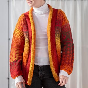 Op de afbeelding: Een felgekleurd, open vest in tinten oranje, rood en geel. De trui heeft een geometrisch patroon op de mouwen en een contrasterende gele bies. Het model draagt een witte coltrui en een donkere jeans.