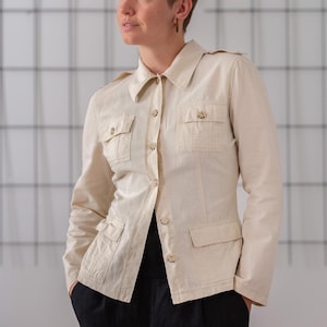 Può includere: Una persona indossa una camicia di lino color crema con colletto e maniche lunghe. La camicia ha due tasche a patta sul petto e una chiusura con bottoni sul davanti. La persona indossa pantaloni neri.