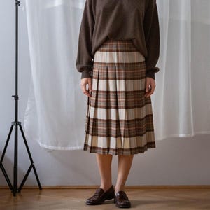 May include: A woman wearing a brown sweater and a plaid skirt. The skirt features a pleated design with brown, beige, and white stripes. She is also wearing brown loafers. The background includes a white curtain and a black tripod.