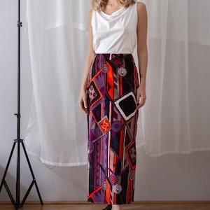 Op de afbeelding: Een vrouw draagt een witte mouwloze top en een lange rok met een patroon. De rok heeft een geometrisch ontwerp in paars, oranje en zwart. Ze draagt zwarte sandalen. De achtergrond bevat een wit gordijn en een zwart statief.