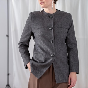 Pode incluir: Um blazer cinza-carvão em mistura de lã com decote redondo sem colarinho, mangas compridas e fecho frontal com quatro botões escuros. O casaco inclui dois bolsos horizontais com debrum no peito e dois bolsos laterais.