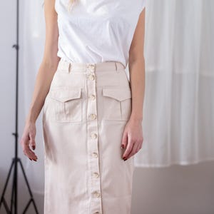 May include: A white sleeveless top with a draped neckline paired with a beige midi-length skirt. The skirt features a front button-down closure, a high waistband with belt loops, and two flap pockets on the front hips, constructed from a solid fabric.