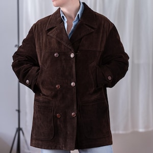 May include: A dark brown suede jacket featuring a double-breasted front with six buttons, a wide collar, and two large patch pockets at the hips. The jacket has long sleeves with buttoned cuffs and is worn over a light blue collared shirt.