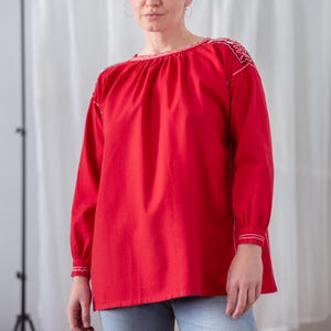 Puede incluir: Una blusa roja de manga larga con detalles bordados en blanco en los hombros y puños. La blusa tiene un escote fruncido y se combina con vaqueros azul claro. El estilo es informal y cómodo.
