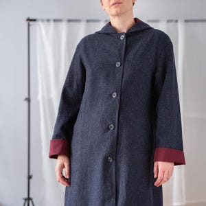 May include: A long, dark blue coat with a hood and button closure. The coat features burgundy cuffs and a burgundy lining in the hood. The coat is knee-length and appears to be made of a textured fabric.