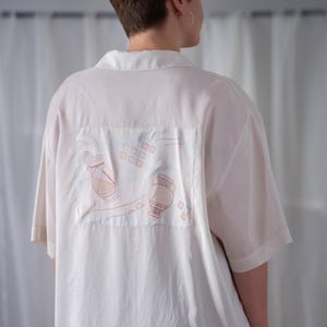 Könnte beinhalten: Cremefarbenes kurzärmeliges Hemd mit Kragen. Auf dem Rücken befindet sich ein rechteckiger Aufnäher mit einer Zeichnung von zwei Vasen und geometrischen Formen in Rosa und Braun. Das Hemd ist aus leichtem Stoff gefertigt.