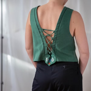 Può includere: Top senza maniche verde con schiena allacciata e applicazione a forma di cuore. Il top ha cuciture nere lungo i bordi e un motivo floreale ricamato sul dettaglio a forma di cuore. La persona indossa pantaloni neri.