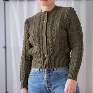 Op de afbeelding: Olijfgroen vest met een ronde hals, lange mouwen en een taille met trekkoord. De trui heeft een kabelgebreid patroon en is gesloten met zilverkleurige knopen. Het model draagt een blauwe jeans.