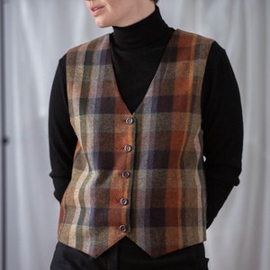 May include: A brown, black, and orange plaid vest with a v-neck and button closure. The vest is worn over a black turtleneck shirt.
