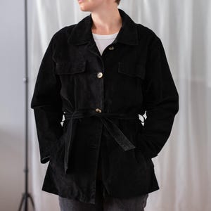 May include: Black corduroy jacket with a belted waist and two front pockets. The jacket has a collared neckline and button closures. The model is wearing a white t-shirt underneath. The jacket is a dark, solid color.
