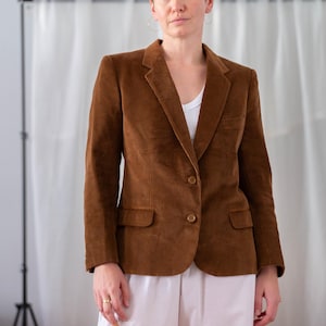 May include: A brown corduroy blazer with a notched lapel and two-button closure. The blazer has two front pockets and is worn over a white top and white trousers. Measurements are not visible in the image.
