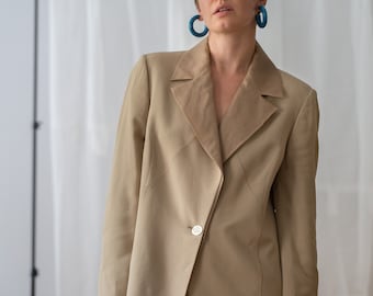 May include: A tan blazer with a contrasting light brown lapel and a single white button closure. The blazer is paired with cream-colored pants. The model is wearing large blue hoop earrings. The background is a white curtain.