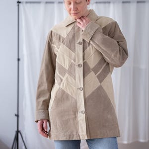 Puede incluir: Chaqueta de ante color beige con diseño geométrico. La chaqueta tiene cuello, mangas largas y cierre de botones. La chaqueta se lleva con vaqueros azules.