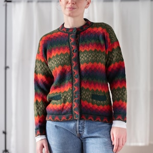 Könnte beinhalten: Bunte Strickjacke mit Knöpfen und rundem Ausschnitt. Der Pullover hat ein Muster aus roten, orangefarbenen, grünen und blauen horizontalen Streifen und Zickzacklinien. Er hat zwei Vordertaschen und wird mit einer blauen Jeans und einem weißen Hemd getragen.