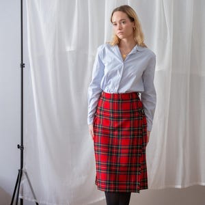 May include: A light blue shirt and a red tartan skirt. The skirt has a wrap design with a buckle fastening. The outfit is completed with black tights and shoes. The style is classic. The skirt's length is approximately 60 cm.