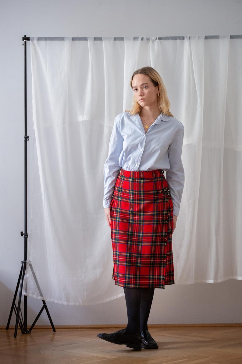 May include: A light blue shirt and a red tartan skirt. The skirt has a wrap design with a buckle fastening. The outfit is completed with black tights and shoes. The style is classic. The skirt's length is approximately 60 cm.