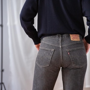 Puede incluir: Vaqueros de mezclilla gris oscuro con un diseño clásico de cinco bolsillos. Los vaqueros tienen un corte de pierna recta y una etiqueta roja de Levi's en el bolsillo trasero. La persona lleva un jersey azul marino.