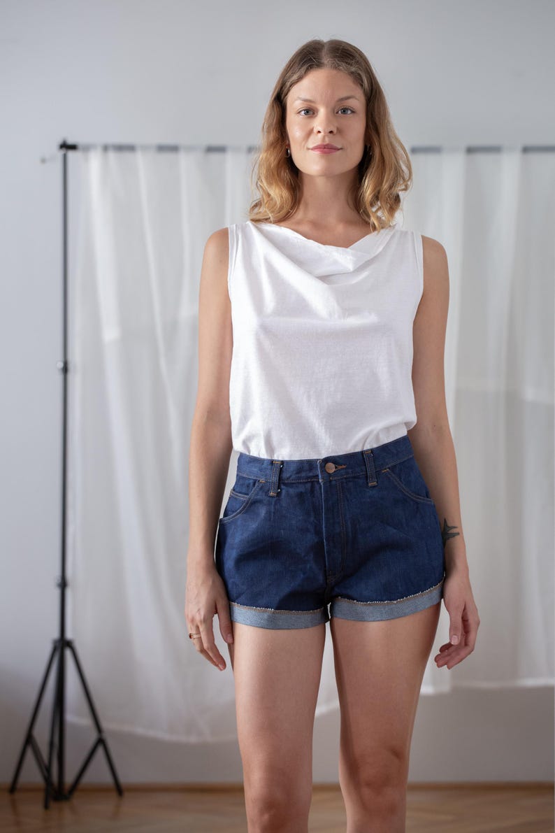 May include: A sleeveless white top with a cowl neckline paired with dark blue denim shorts. The shorts have a rolled hem and classic five-pocket styling. The outfit is simple and casual.