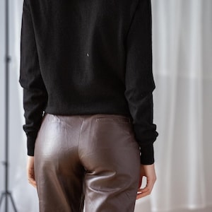 May include: A black long-sleeve sweater paired with brown leather pants. The pants have a slight sheen and a tailored fit. The model's back is facing the camera, showcasing the outfit's silhouette.