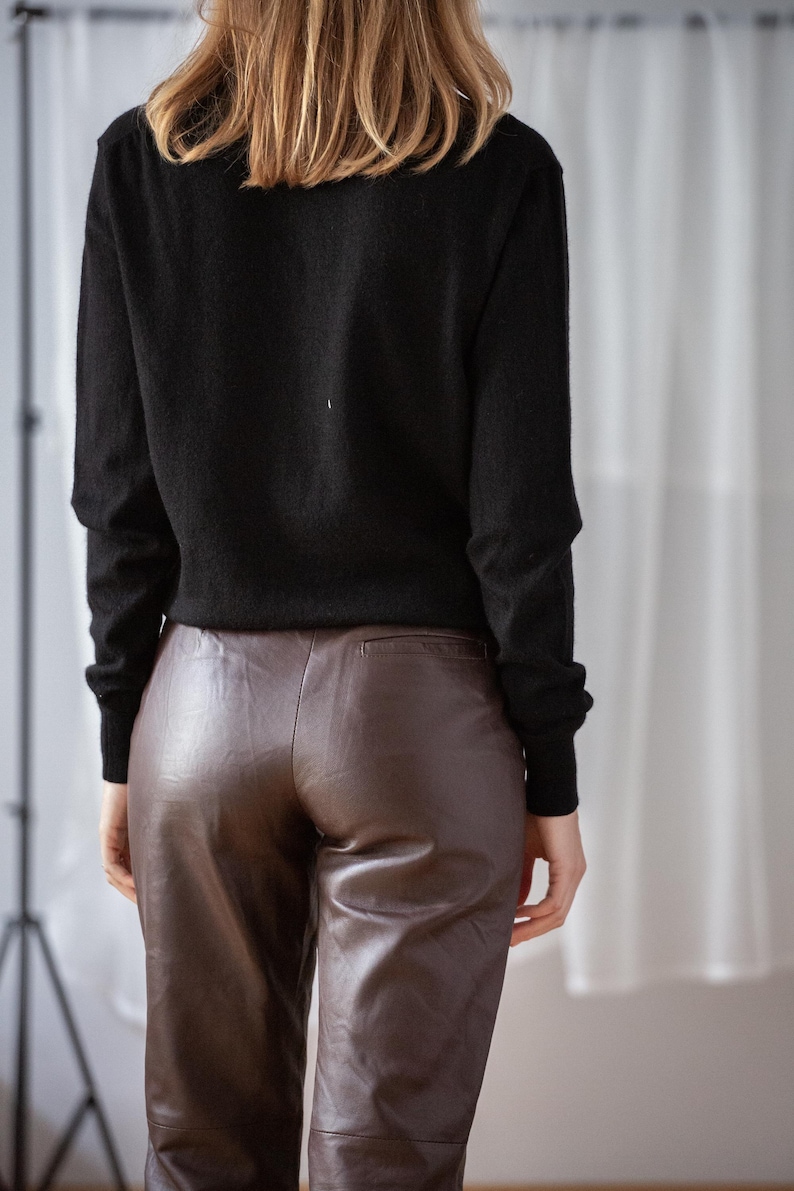 May include: A black long-sleeve sweater paired with brown leather pants. The pants have a slight sheen and a tailored fit. The model's back is facing the camera, showcasing the outfit's silhouette.