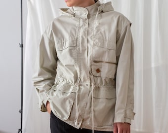 Puede incluir: Una chaqueta con capucha de color beige claro con cintura de cordón y varios bolsillos. La chaqueta está hecha de una tela ligera y tiene un corte holgado.