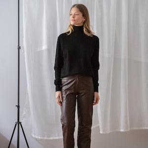 May include: A person wearing a black turtleneck sweater and brown leather pants. The outfit is paired with black sandals. The background includes a white curtain and a black tripod.