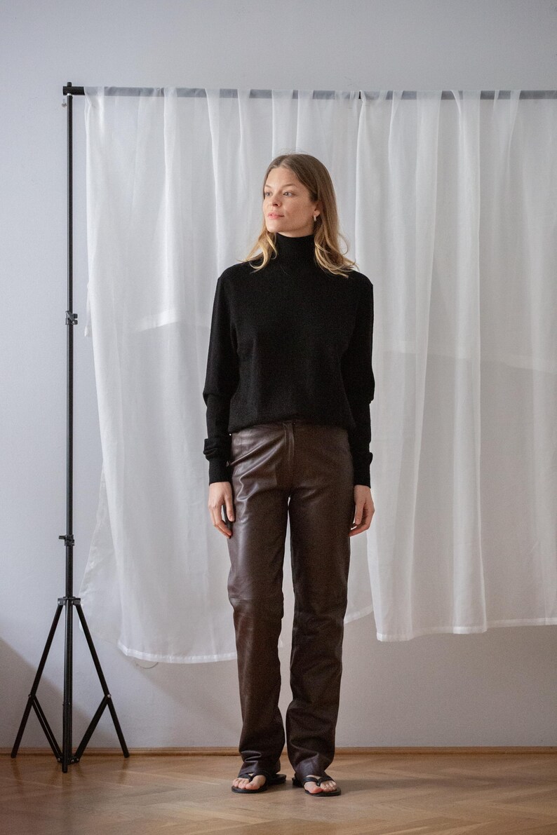 May include: A person wearing a black turtleneck sweater and brown leather pants. The outfit is paired with black sandals. The background includes a white curtain and a black tripod.