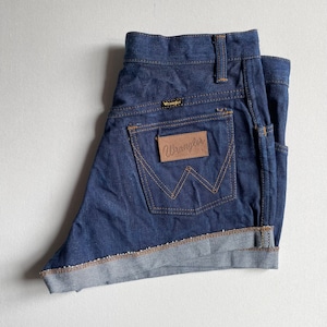 May include: Dark blue denim Wrangler shorts with a folded hem. The shorts feature the Wrangler logo on the back pocket and a small logo above the back pocket. The stitching is a contrasting tan color.